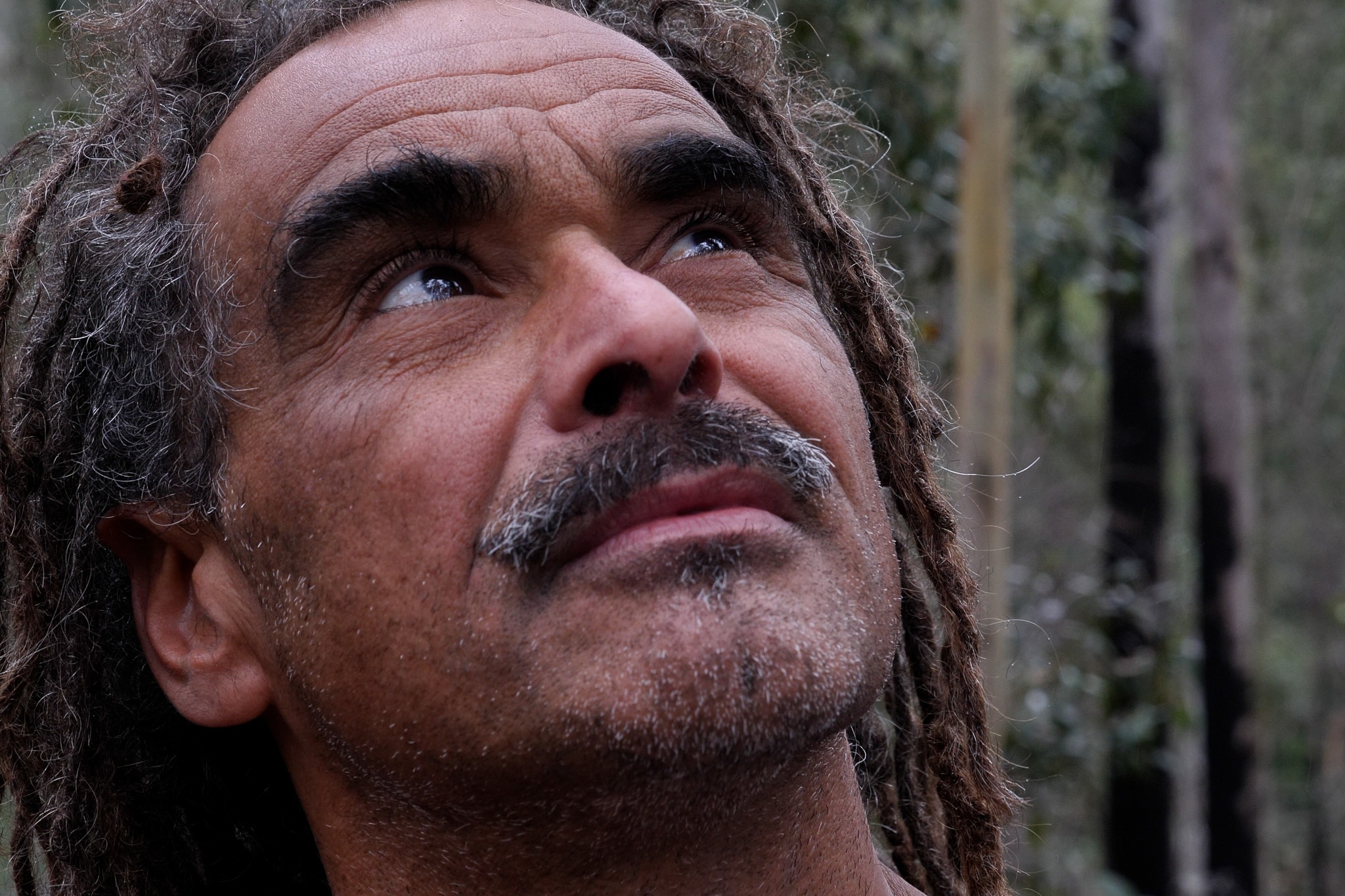 Close up of man looking up at trees with serious expression, has dregs and a grey and black moustache.