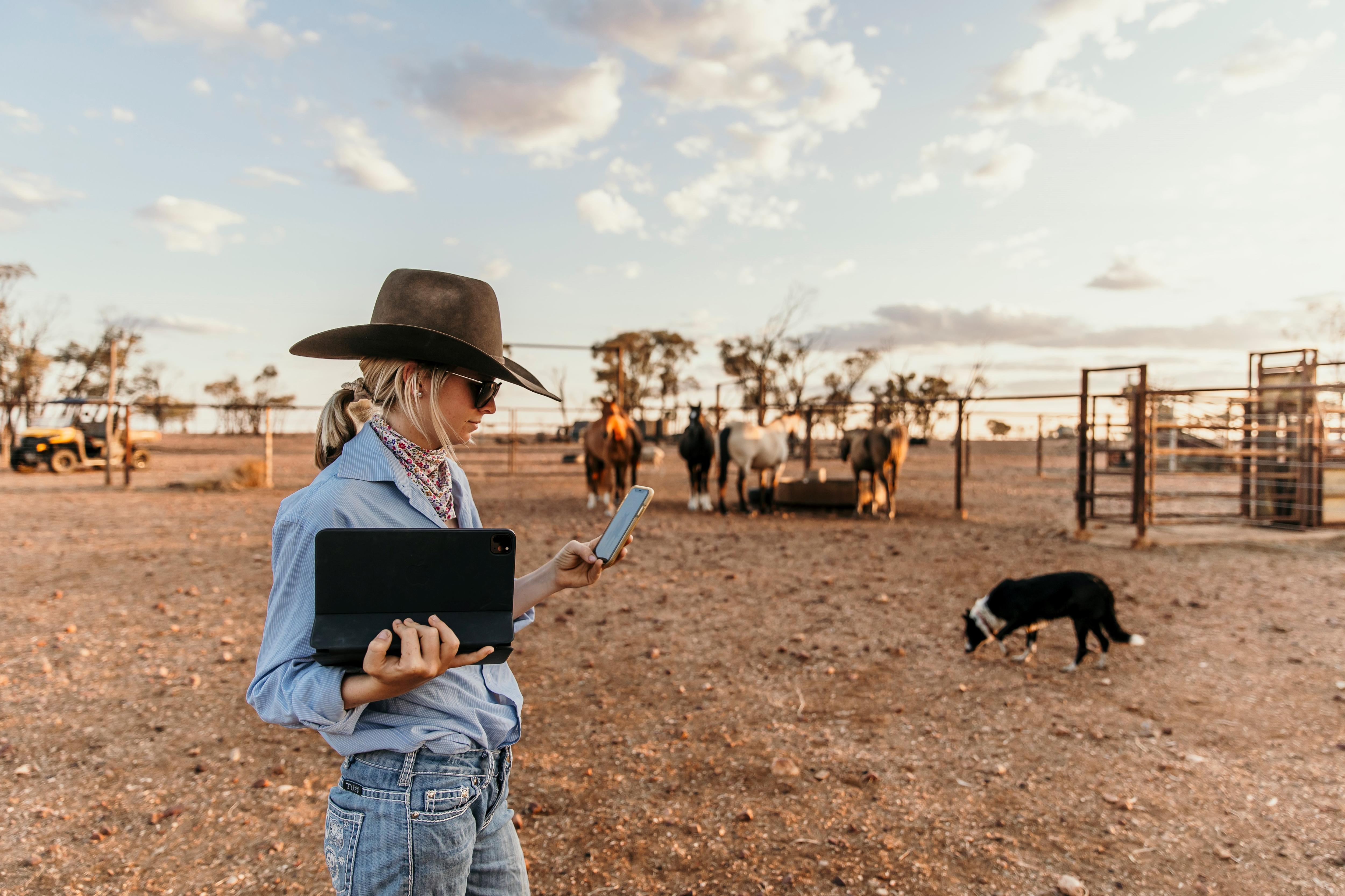 A woman in a blue shirt and akubra holds a laptop and phone in a yard with horses.