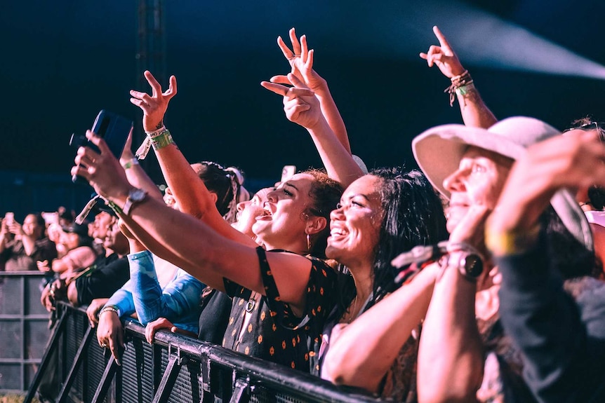 Women in the front row of a concert grin at the stage and hold their hands up