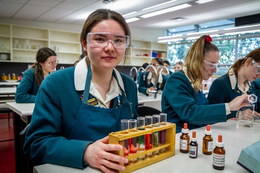 Amelie wears goggles in a school science lab, holding a stand containing test tubes with different coloured fluids inside them.