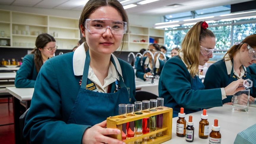 Amelie wears goggles in a school science lab, holding a stand containing test tubes with different coloured fluids inside them.