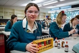 Amelia wears goggles in a school science lab, holding a stand containing test tubes with different coloured fluids inside them.