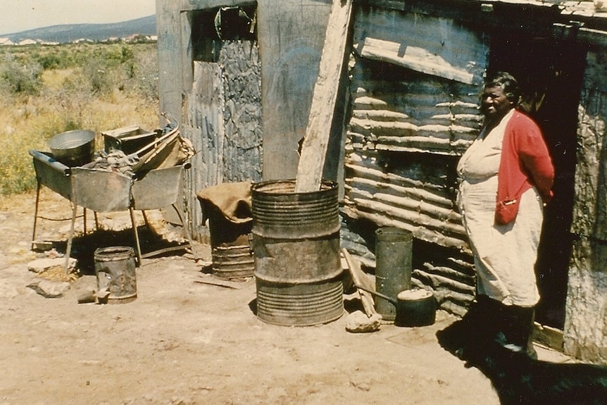 Indigenous woman standing on right in front of corrugated iron make-shift shed with dog 