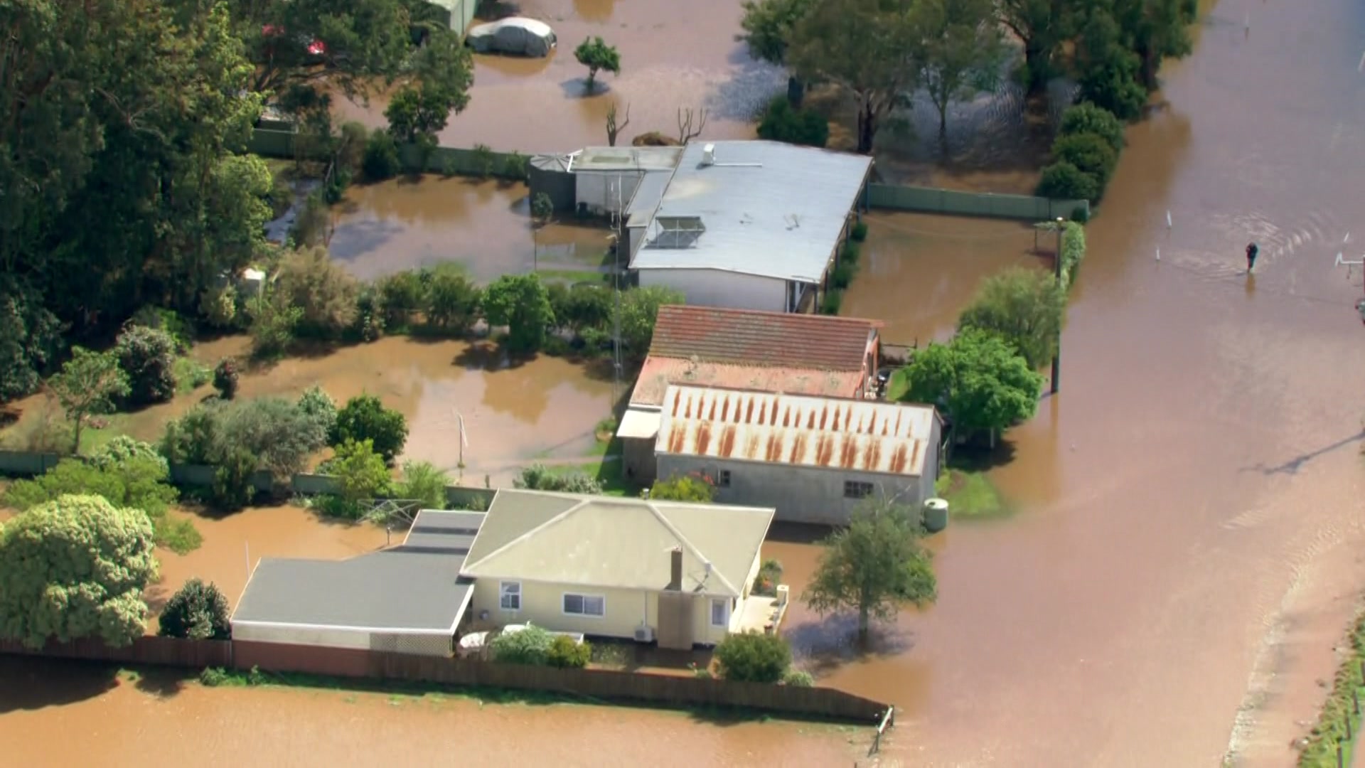 An aerial shot of brown murky flood waters surrounding several properties and a person walking through the water