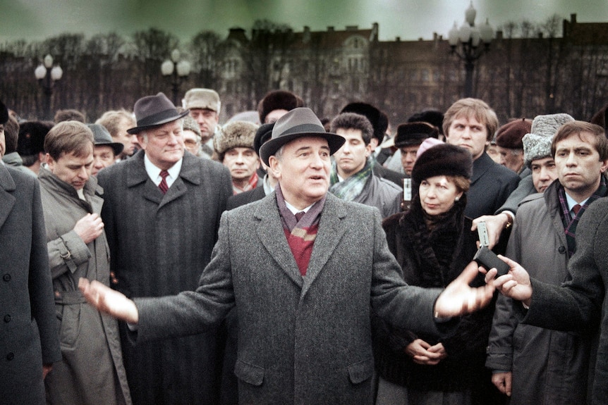 A man (Mr Gorbachev) stands at the foreground of a mass of people, gesturing with his hands while he speaks. 