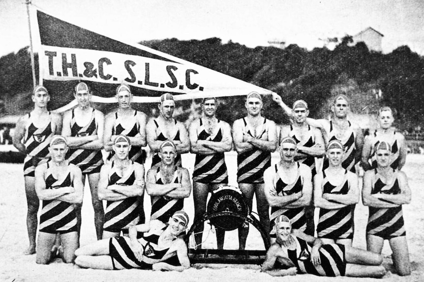 Male surf lifesavers standing for a group shot with their flag on the beach.