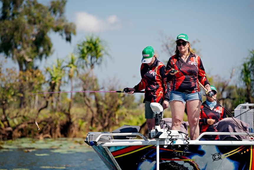 Three women stand in a boat with fishing rods, one at the helm controlling steering