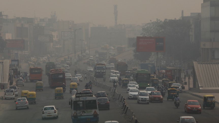 Cars and motorcycles drive along a wide road full of heavy smog in an Indian city.