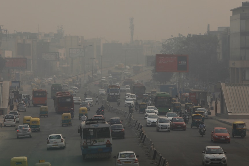 Cars and motorcycles drive along a wide road full of heavy smog in an Indian city.