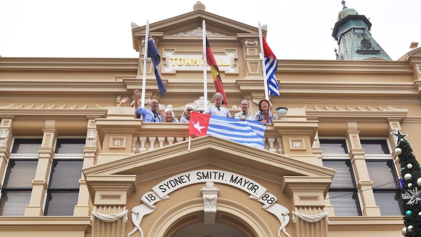 A group of people in balcony in a building with flags of Australia, indigenous Australians, and Papuan.
