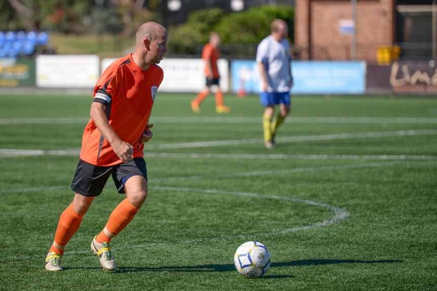 A man in an orange shirt plays football, a soccer ball is in front of him. 