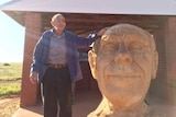 A man stands next to a large stone-carved head of his own likeness.