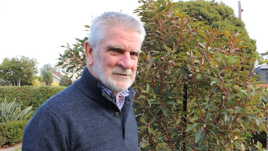 An older, grey-haired and bearded man in a blue jumper stands in front of a tree.