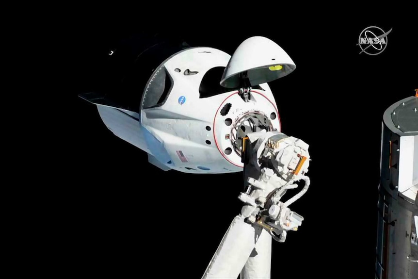 A white triangular space capsule docks onto two white arms of the International Space Station.