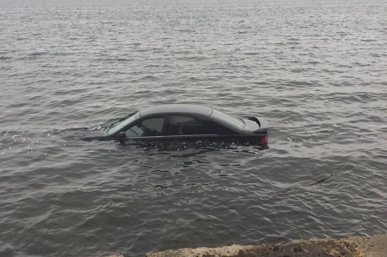 A black car lies partially submerged in the Swan River near the shore.