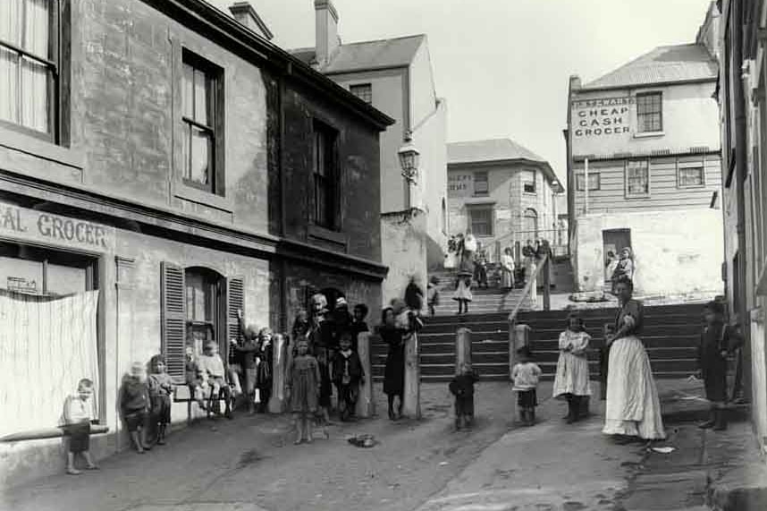 Black and white photo from 1901 of families in the street, in front of two grocers.