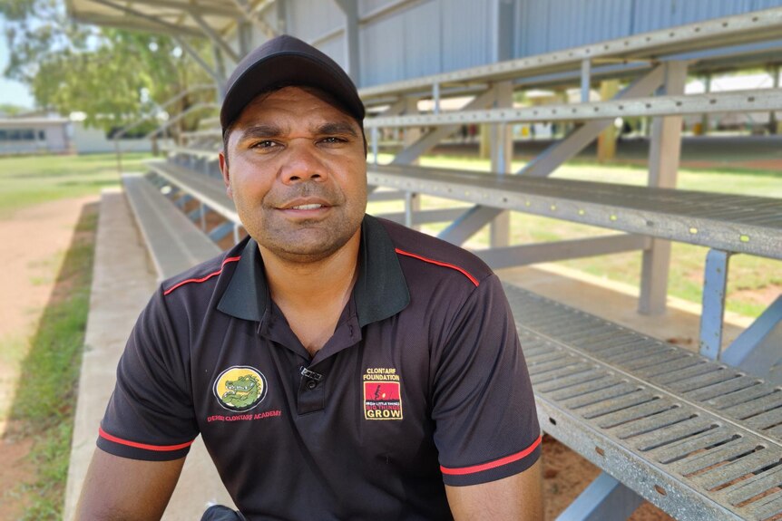 An Indigenous man sits on a spectator stand next to a football oval