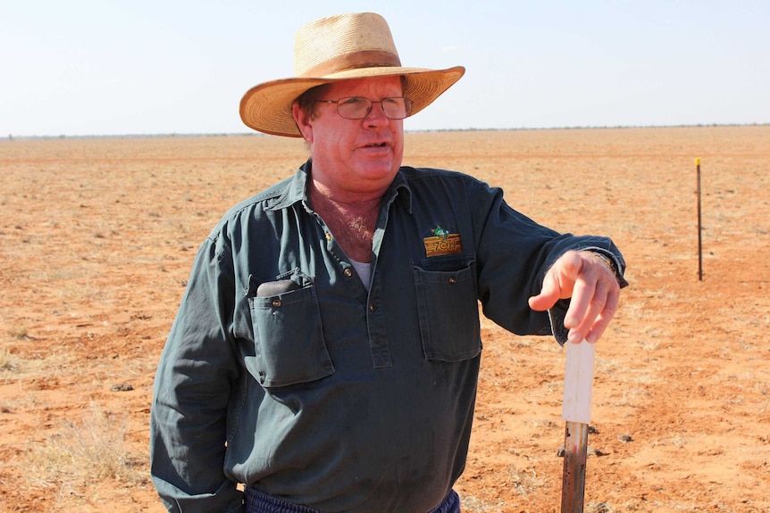 A man wearing a hat leans again a post as he looks out on a parched landscape.