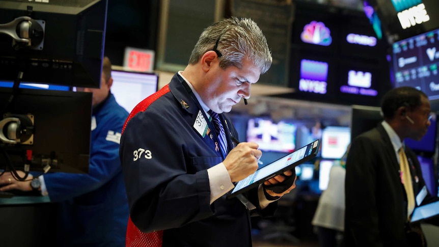 A stock trader looks down at a tablet as he works on the floor of a busy stock exchange while surrounded by screens.