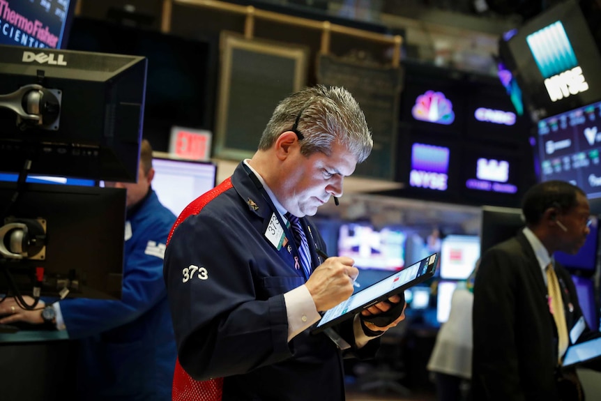 A stock trader looks down at a tablet as he works on the floor of a busy stock exchange while surrounded by screens.
