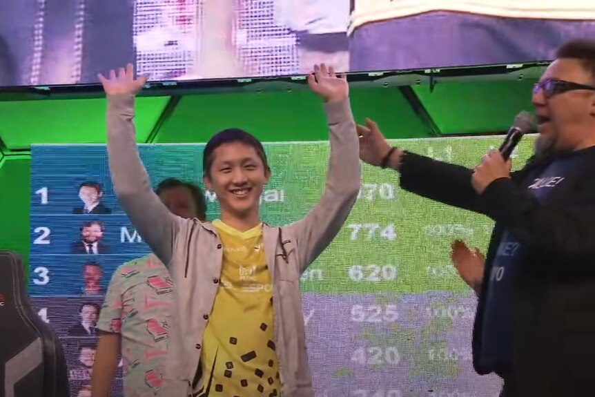A medium shot of a man of Asian descent smiling and raising his hands in the air on stage at an e-sports event