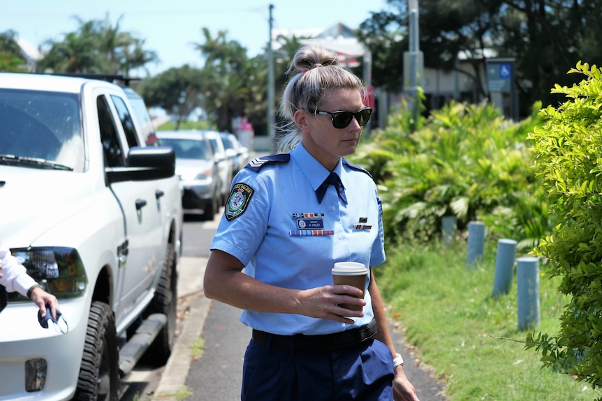 A blonde woman in blue police uniform with sunglasses outside holding coffee cup