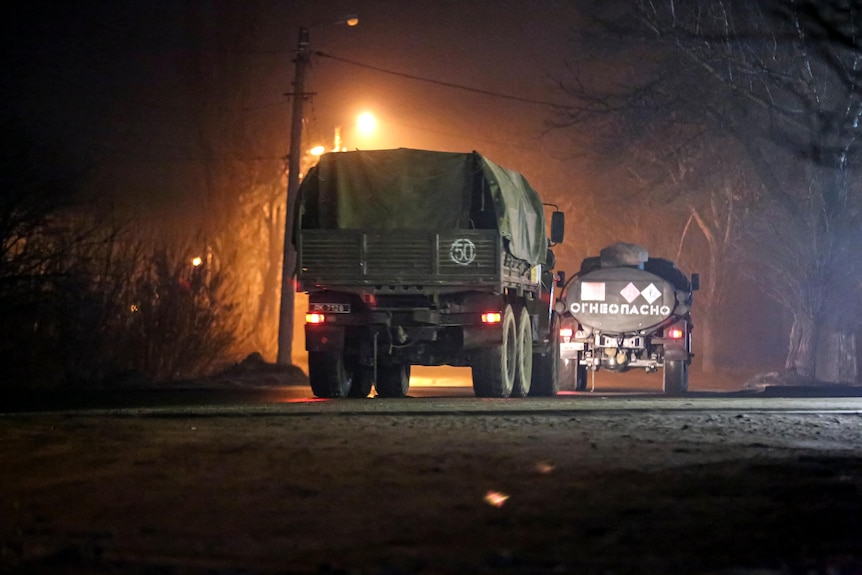 A convoy of military trucks drives along a road at night. 