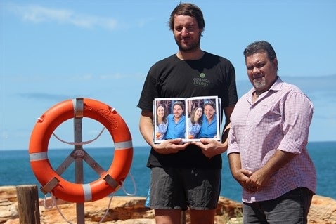 A tall man stands holding a photobook of him with a smiling woman, beside an Aboriginal man and a life ring.