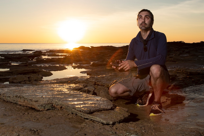 Dr Romilio in the field in Broome, crouched on rocks near ocean sunset behind