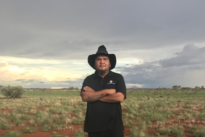 A man in black polo t-shirt wears cowboy hat and stands with arms crossed on cloudy day in flat bushy red dirt Pilbara terrain.