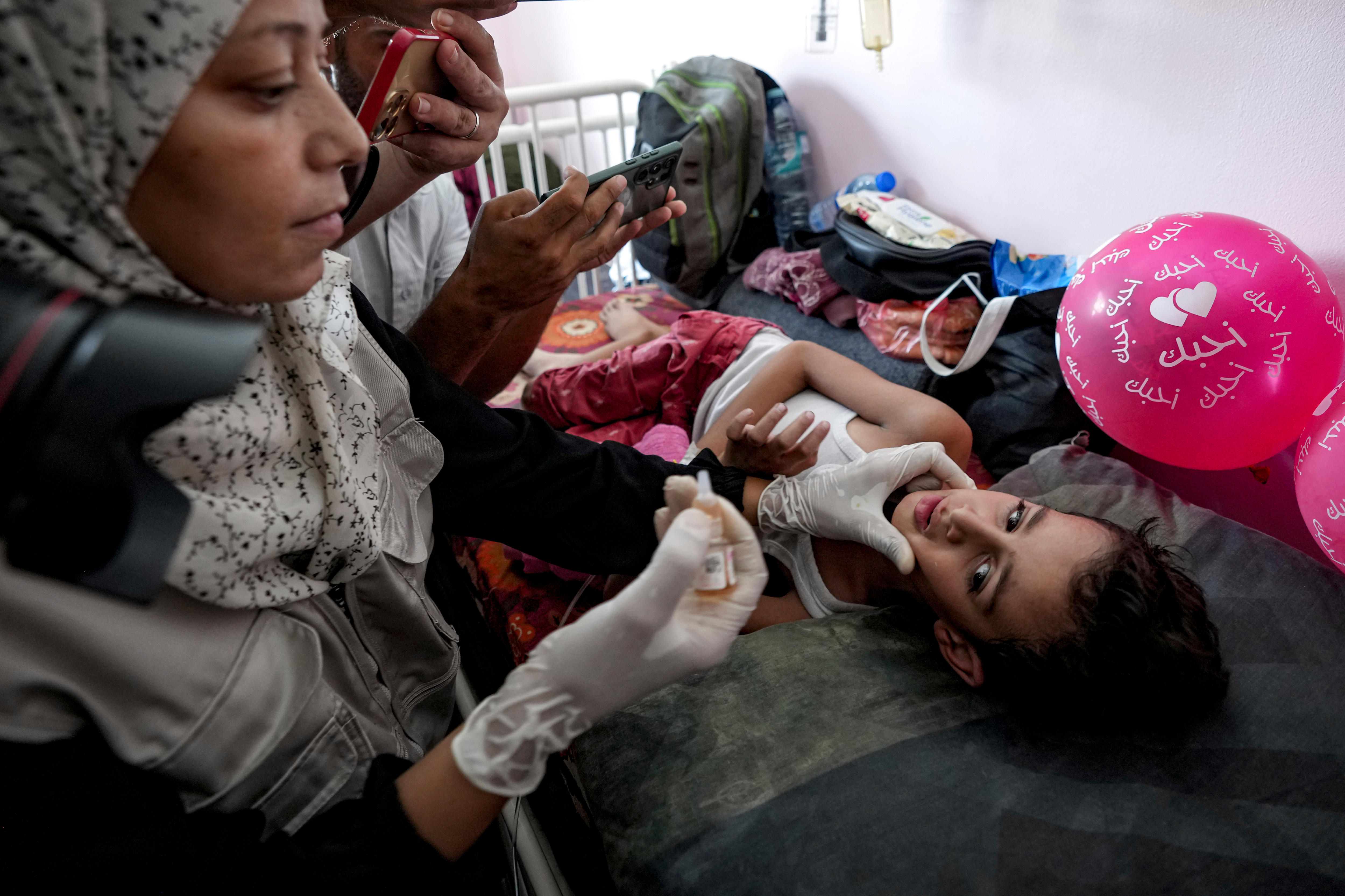 A middle-aged woman readies a small dripper of liquid for a small child lying on a bed next to some pink balloons.