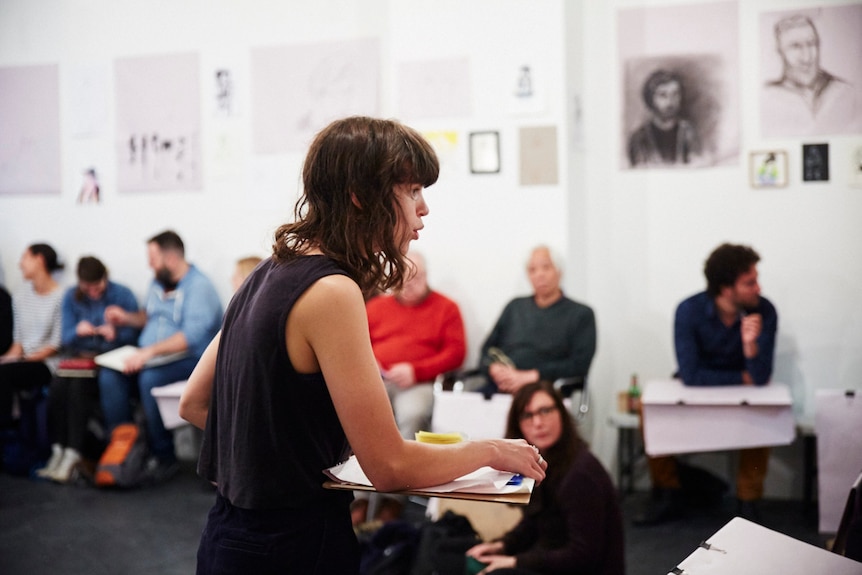 Louise Klerks stands in the foreground, while people sit behind her with sketch pads.