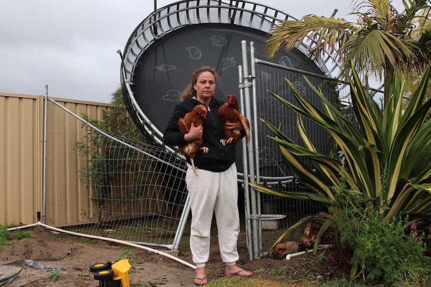 A woman stands in her back yard holding two chickens in front of a trampoline that has been thrown into the garden and fence.
