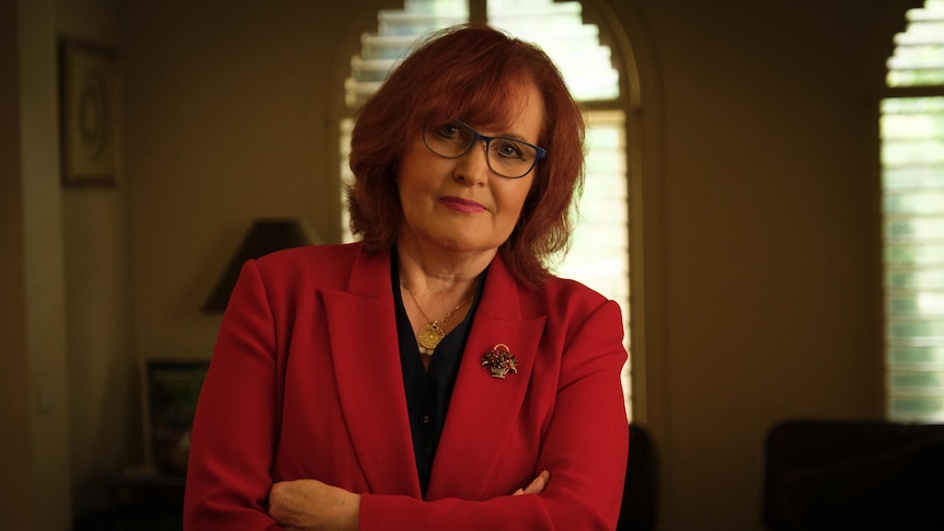 Karen wears a red blazer and stands with her arms crossed with a serious expression in a living room framed by an arched window.