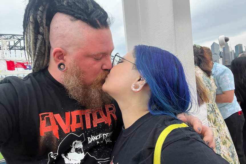 A man with his head shaved on the sides kisses a woman with blue hair.