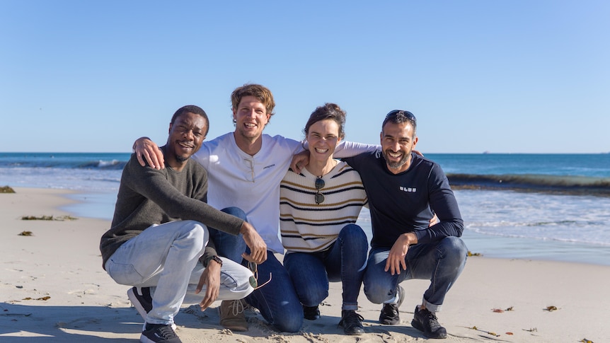 Four smiling people kneel on a sunny beach, casually dressed, their arms around each others' shoulders. 