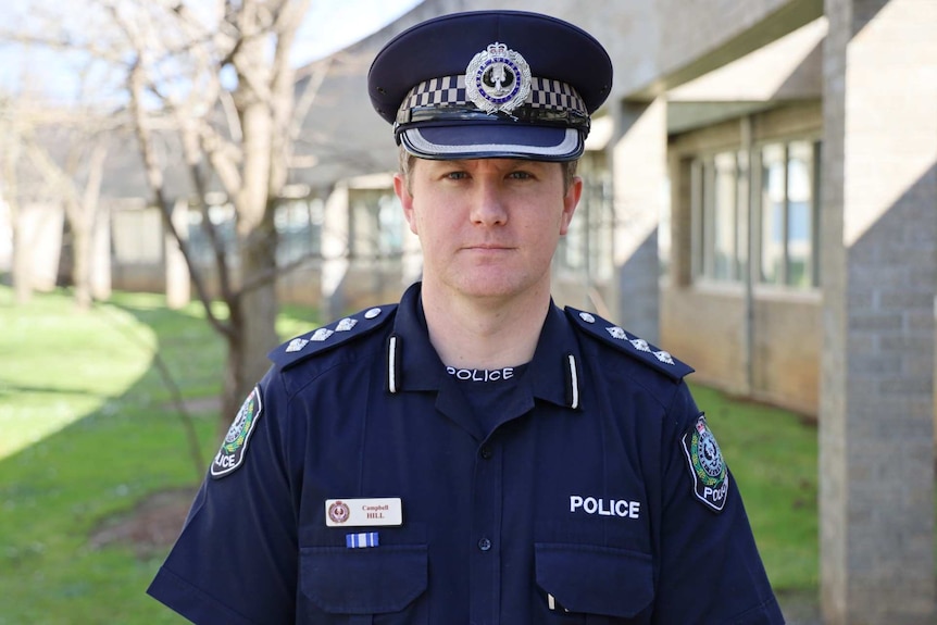 A man stands in front of a grey building, wearing a navy blue South Australian police uniform and hat.