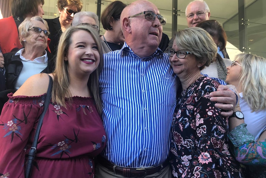 Peter Creigh, in a striped blue shirt, is hugged by two women.