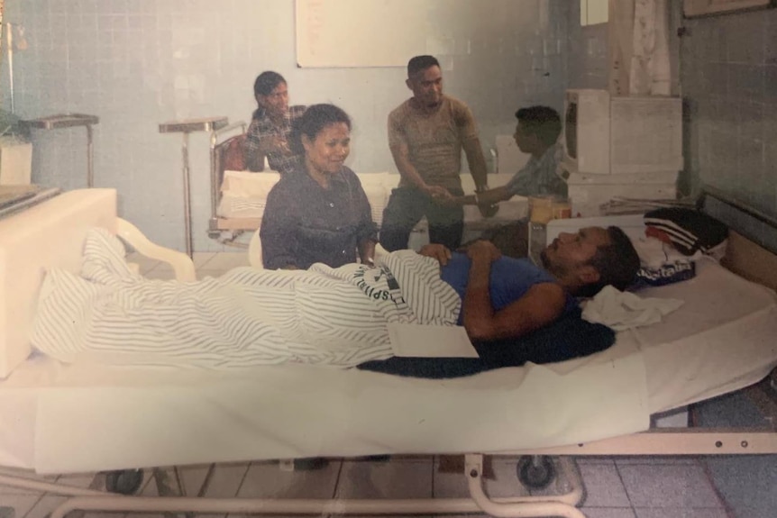 A man lies in a hospital bed while his loved ones sit around him.