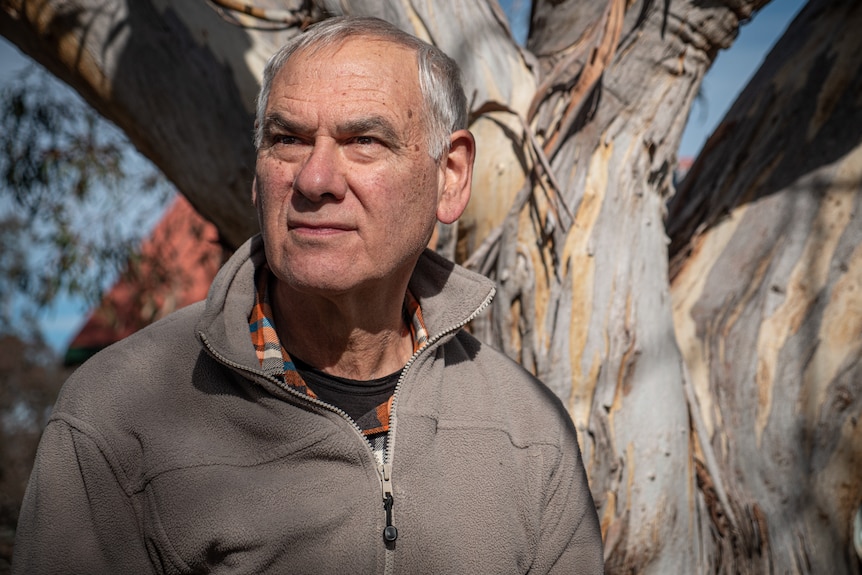 A man wearing a flannelette and polar fleece sits in front of a gum tree staring out into the distance..