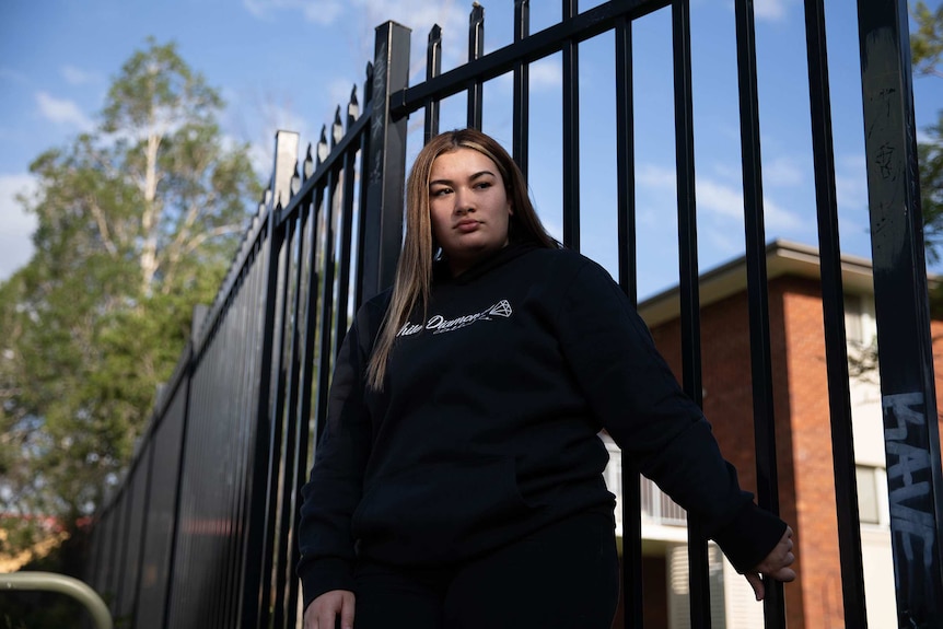 Manifold rapper Moana leaning against a black metal fence, with red-brick building in background.
