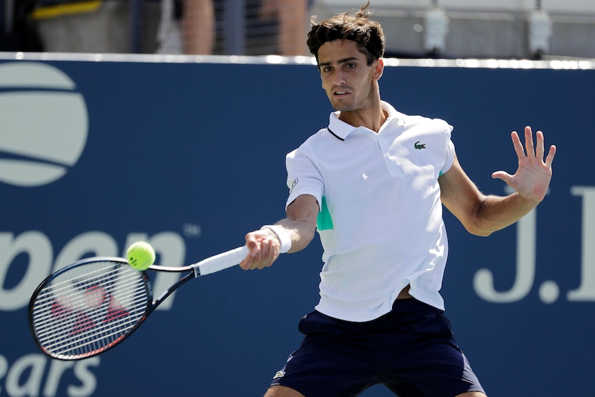 A male tennis player in a white polo shirt hits a forehand
