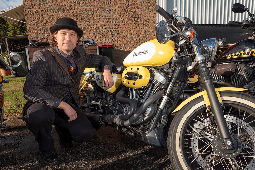 A man in a suit and bowler hat crouched next to his motorcycle.