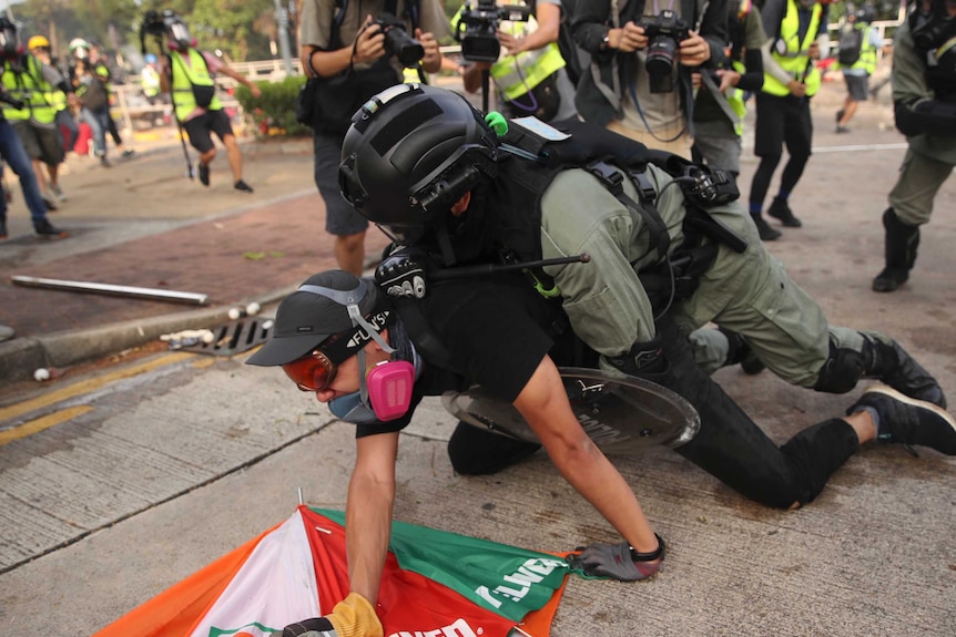 A police officer wearing riot gear is holding a student to the ground as cameramen and other police surround them.