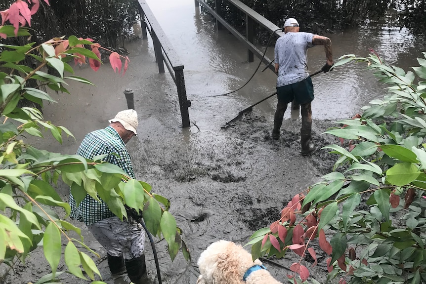 Two men in gum boots rake a thick layer of mud off a grassed area of a garden.