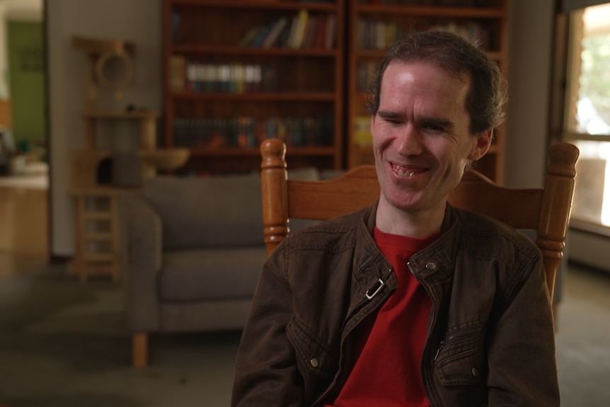 Mick Curran sits in a dining chair in a loungeroom. He wears a red shirt and brown jacket and is smiling