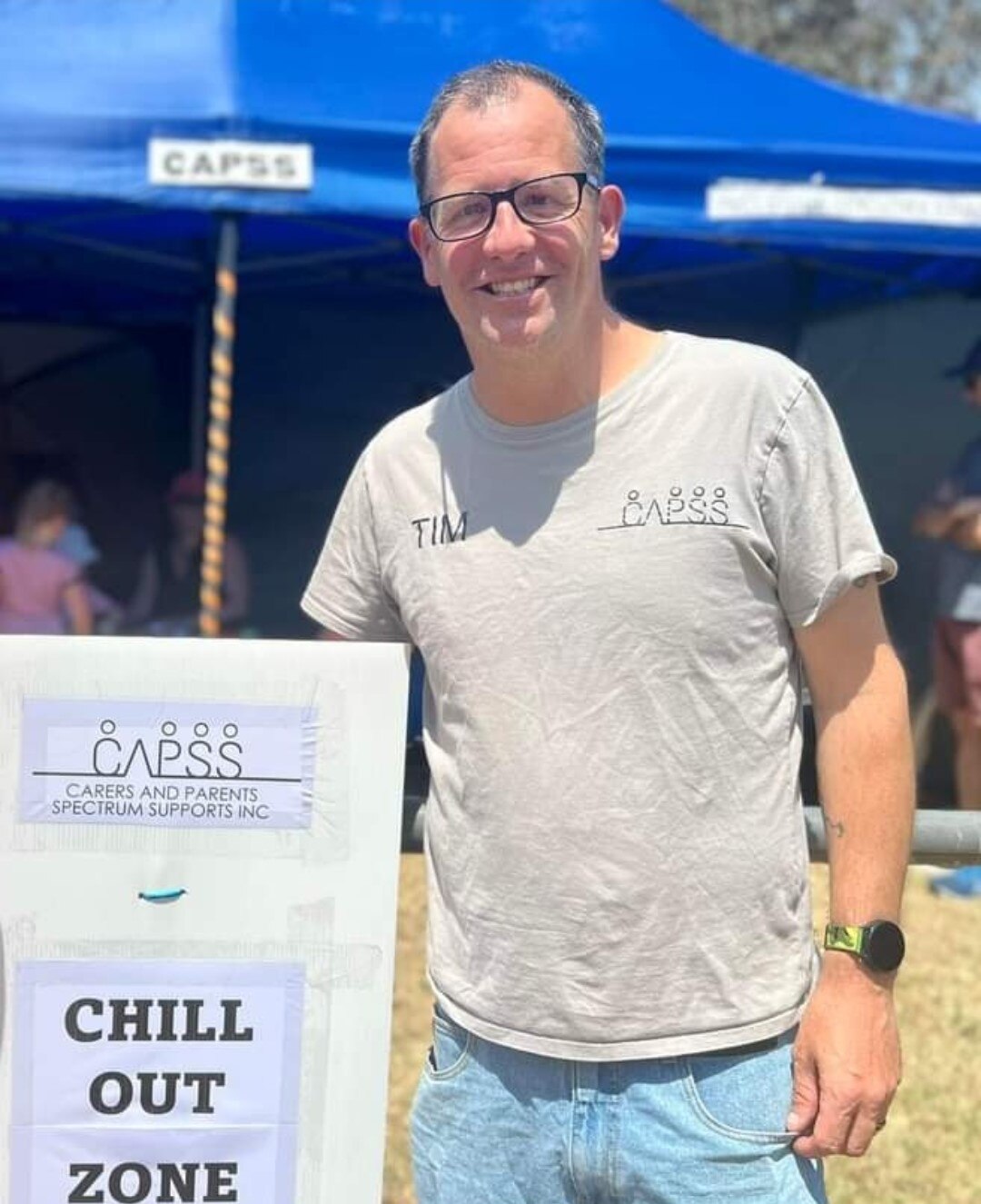 A man in glasses, grey t-shirt and blue jeans standing outside beside a sign that says CAPSS chill out zone.