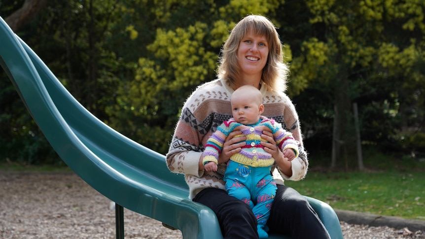 A woman sits on the end of a playground slide with a baby on her lap