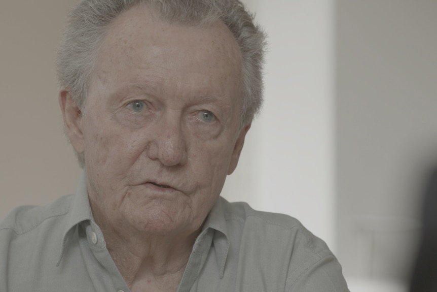 An older man with blue eyes and grey hair talking to someone sitting opposite.