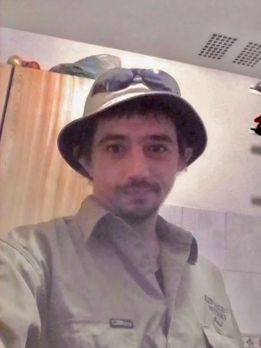 A young man with a light beard in a bucket hat stands indoors, taking a selfie.